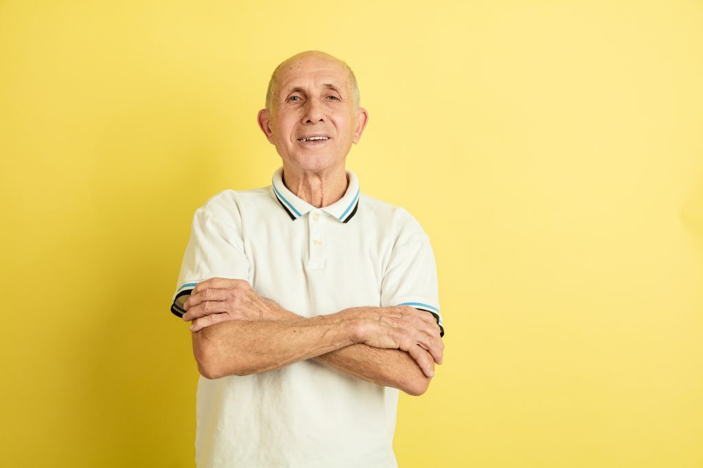 Hands crossed. Caucasian senior man's portrait isolated on yellow studio background. Beautiful male emotional model. Concept of human emotions, facial expression, sales, wellbeing, ad. Copyspace.